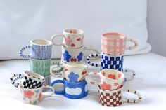 a pile of coffee mugs sitting on top of a white bed covered in pillows