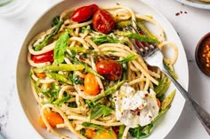 a white bowl filled with pasta, tomatoes and asparagus on top of a table