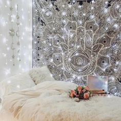 a white bed sitting in front of a wall covered in lights and blankets next to a vase filled with flowers