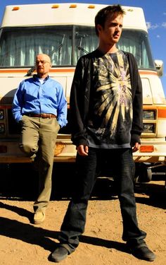 two men standing next to each other in front of a bus on dirt ground with blue sky and clouds behind them