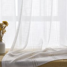 a white vase with flowers in it sitting on a table next to a sheer curtain