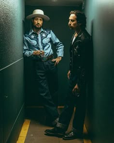 two men standing next to each other in a hallway with one man wearing a cowboy hat