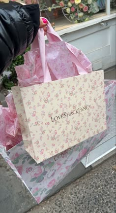 a person is holding two shopping bags that say loveshape fancy on them, in front of a flower shop