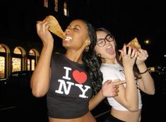 two young women eating pizza outside at night, one is biting into the other's mouth