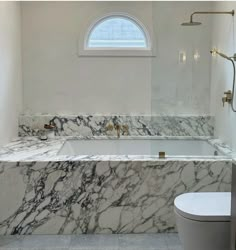 a marble bathtub in a bathroom with a toilet and window above the tub is an arched window