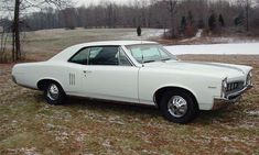 an old white car is parked in the grass near some trees and snow covered ground