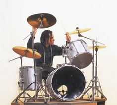 a man with dreadlocks is playing drums on a wooden stand in front of a white wall