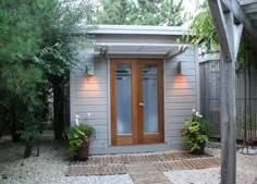 a small gray house with wooden doors and plants in the front yard, surrounded by trees