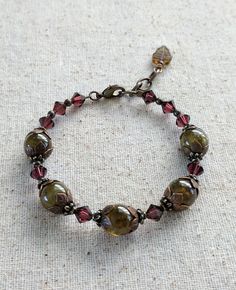 A classy, eye-catching bracelet made with glowing Green Garnet and sparkling, wine-colored crystals. It is accented with decorative floral antiqued brass bead caps and findings. It is finished with a lobster claw clasp and features an extension chain that makes it easy to put on and adjustable. It is available in 2 lengths- 6 3/4"-7 1/2" and 7 1/2" -8". Classy Beaded Bracelets, Beaded Crystal Bracelets, Vintage Red Gemstone Beads Jewelry, Vintage Green Beaded Bracelets, Handmade Antique Green Bracelet, Vintage Beaded Bronze Bracelets, Nature-inspired Brown Jewelry With Gemstone Beads, Antique Accessories