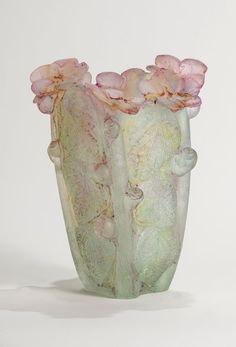 a green vase with pink flowers in it on a white surface and behind it is a light colored background