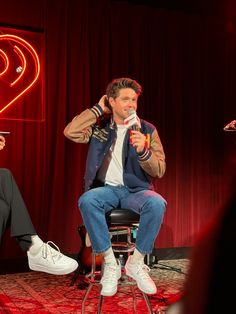 two people sitting on chairs in front of a red curtain and one person holding a microphone