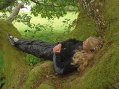 a woman laying on the ground next to a mossy tree trunk with her hair blowing in the wind