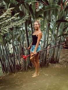 a woman standing in front of some plants holding a red object with one hand and wearing a black tank top