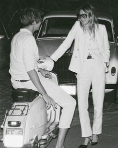 a man and woman standing next to each other on a scooter in front of a car