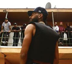 a man wearing a baseball cap and vest standing in front of a balcony with people watching