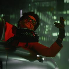 a man in red jacket and sunglasses leaning on the hood of a silver car at night