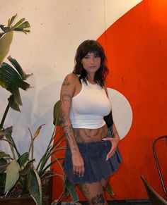 a woman with tattoos standing in front of a wall and potted plant behind her