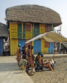 some people are sitting under an umbrella in front of a hut with thatched roof