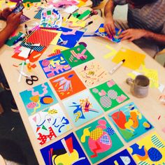 two men sitting at a table making art work on paper with scissors and colored papers