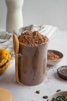 a jar filled with chocolate powder next to two bowls of seeds and banana's