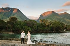 a bride and groom holding hands on the shore of a lake with mountains in the background