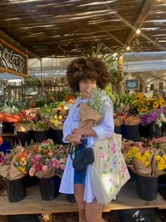 a woman standing in front of a bunch of flowers holding a bag and looking at the camera