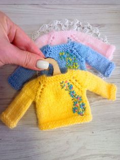 a hand is holding an object in front of three knitted sweaters on a table