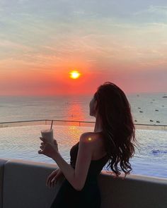 a woman in a black dress is holding a drink and looking at the sunset over an outdoor swimming pool