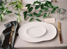 a place setting with white plates and silverware on a linen tablecloth, surrounded by greenery