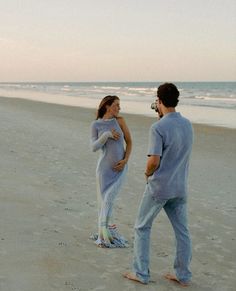 a pregnant woman standing on top of a sandy beach next to a man in blue