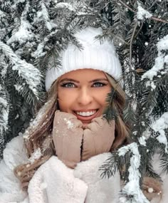 a woman wearing a white hat and scarf in front of snow covered pine trees with her hands on her face