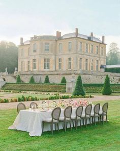 an outdoor dining table set up in front of a large building with hedges and bushes