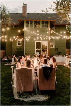 a group of people sitting around a dinner table in front of a house with string lights