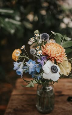 a vase filled with lots of different flowers on top of a wooden table in front of trees