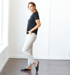 a woman in black shirt and white pants standing on wooden floor next to large window