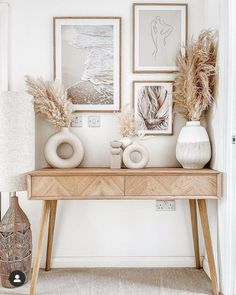 a wooden table topped with two vases filled with dry grass next to pictures on the wall