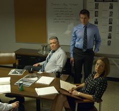 three people sitting at a table with papers and pens in front of them, one person standing