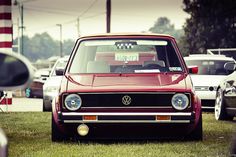 a red car is parked in the grass near other cars and people are looking at it