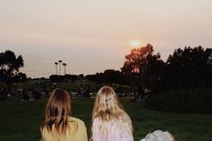 two young women sitting on the grass watching the sun go down over the ocean and trees