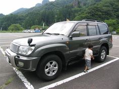 a young boy standing next to a parked car in a parking lot with mountains in the background