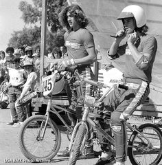 two men riding bikes with helmets on in front of a group of people sitting behind them