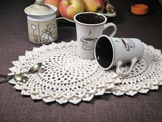 two coffee mugs sitting on top of a doily with spoons and apples in the background