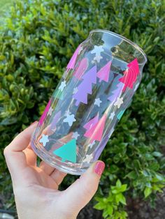 a person holding up a clear cup with colorful stars and trees on the bottom, in front of some bushes