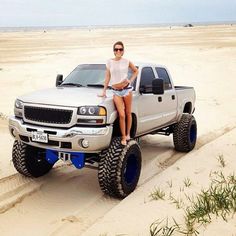 a woman sitting on top of a silver truck in the middle of a sandy area