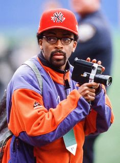 a man in an orange and purple jacket is holding a video camera while wearing a red hat