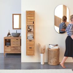 a woman walking past a bathroom sink next to a wooden cabinet and mirror on the wall