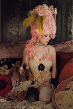 a woman sitting on top of a bed wearing a pink and yellow dress with feathers