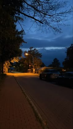 an empty street at night with cars parked on the side and trees lining the road