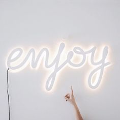 a person pointing to the word enjoy on a wall with a neon sign above it