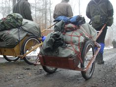 two men pushing a cart with bags on it down a dirt road in the woods